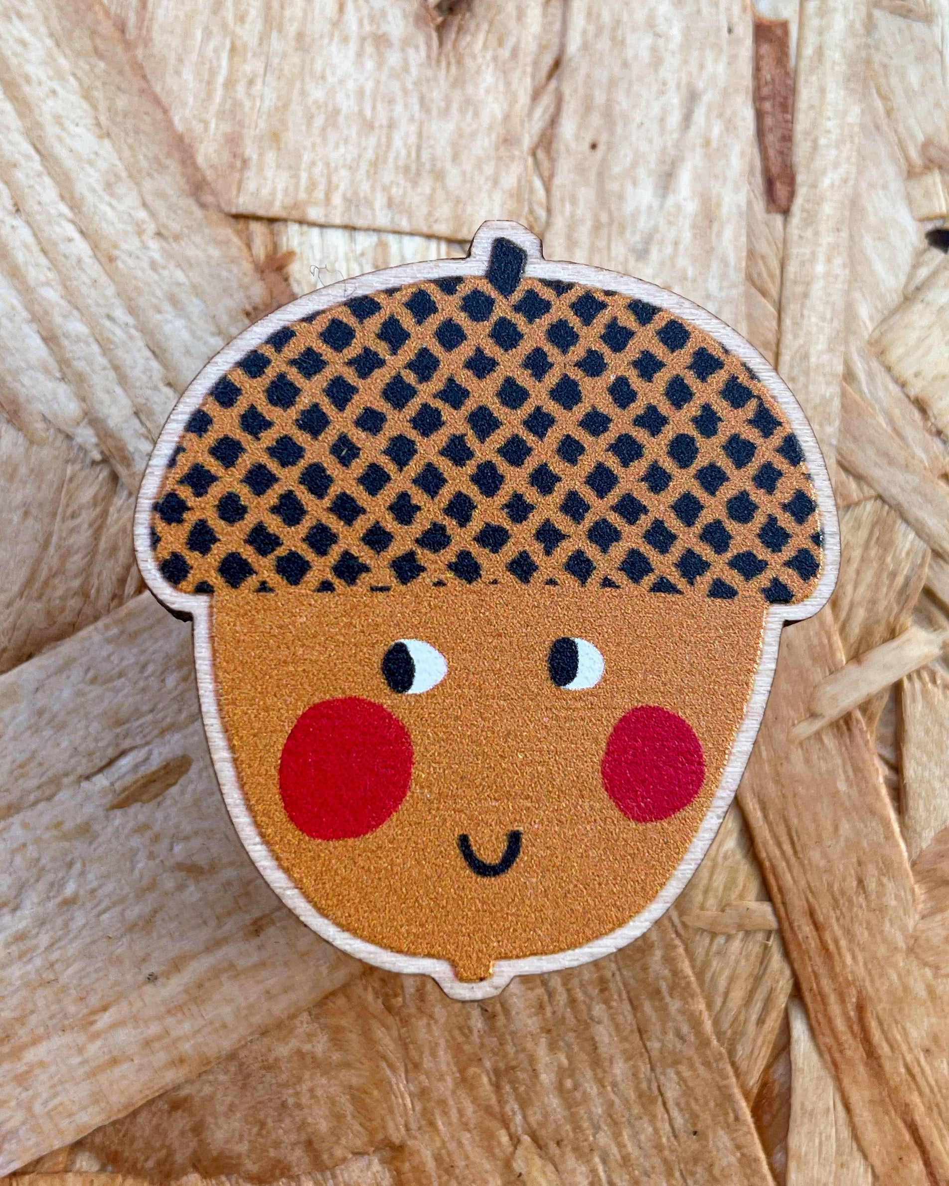 Wooden pin badge - Acorn-shaped character with a checkered cap on a wooden background. 