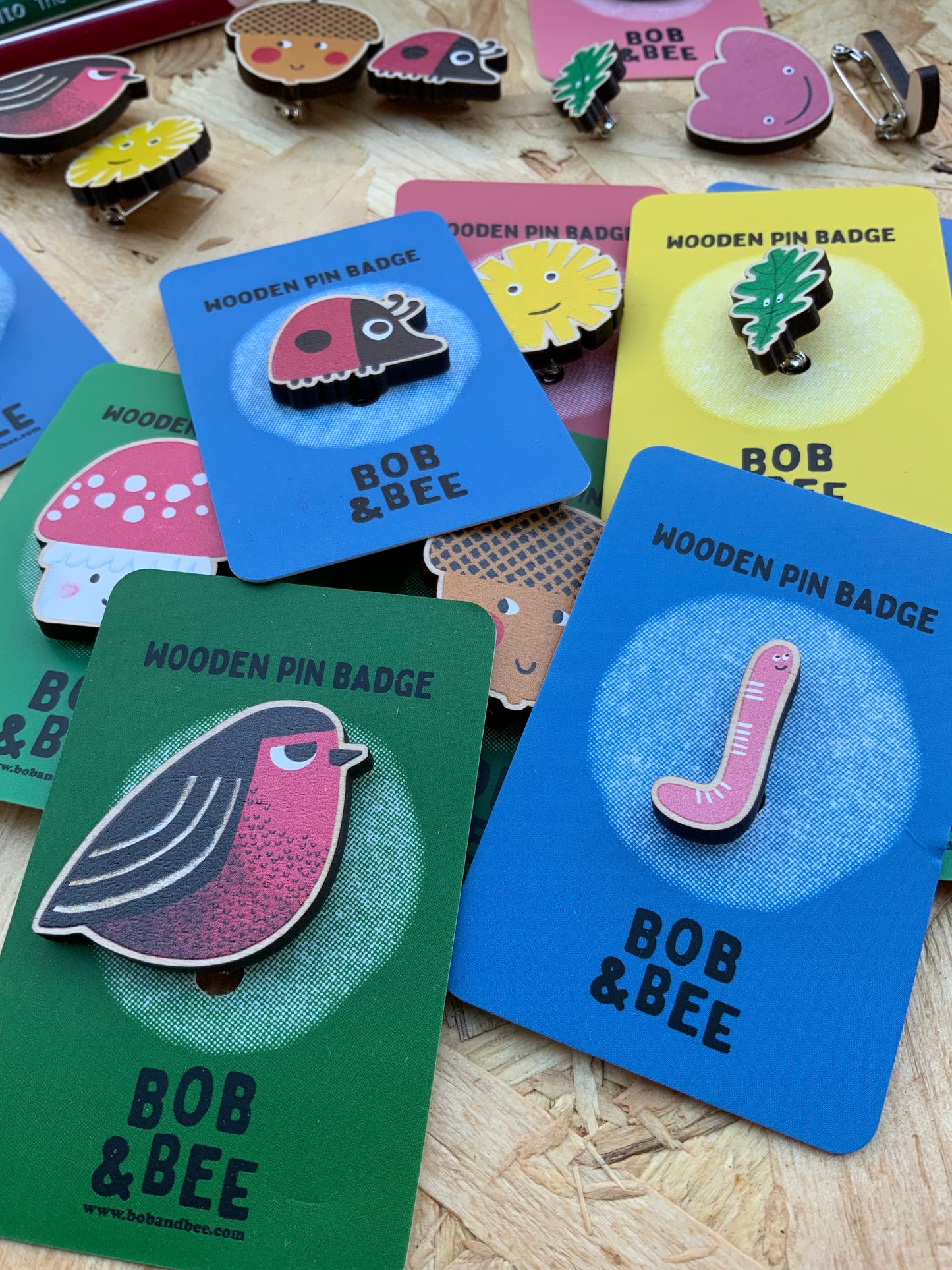 Collection of colourful, fun 'Bob & Bee' wooden pin badges on cards with a wooden surface background. 