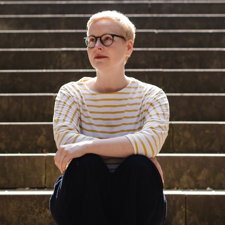 A woman is sitting on stone steps. She has short blonde hair, wears glasses and a stripy t-shirt. She is looking off to the side. 