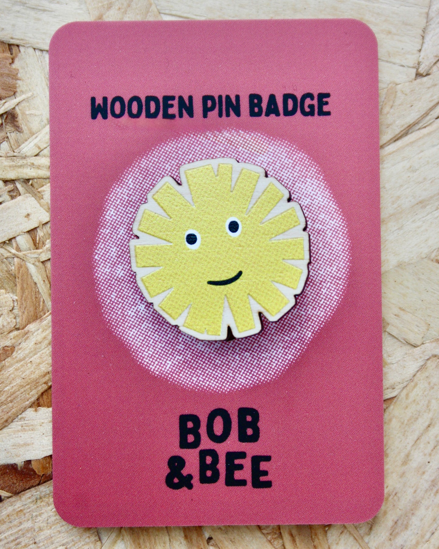 Yellow sun-shaped dandelion wooden pin badge on a pink card with 'BOB & BEE' branding. Wooden background. 
