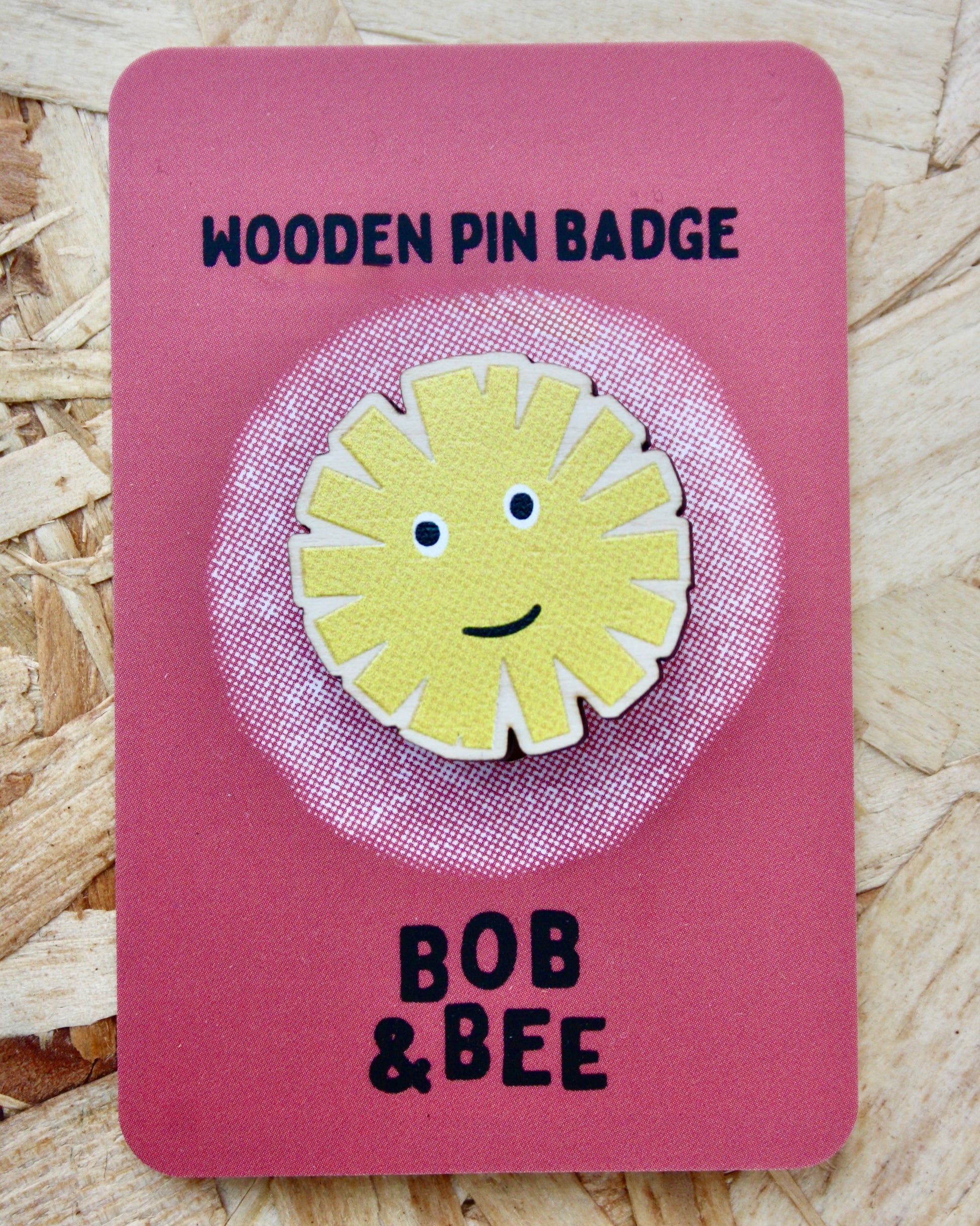 Yellow sun-shaped dandelion wooden pin badge on a pink card with 'BOB & BEE' branding. Wooden background. 
