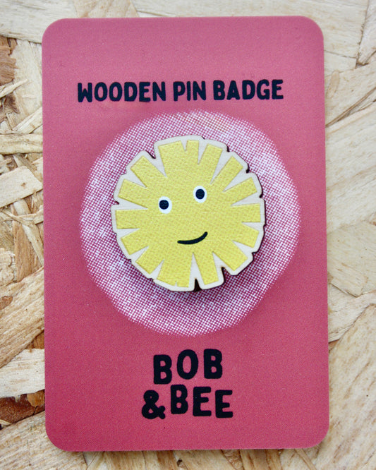 Yellow sun-shaped dandelion wooden pin badge on a pink card with 'BOB & BEE' branding. Wooden background. 